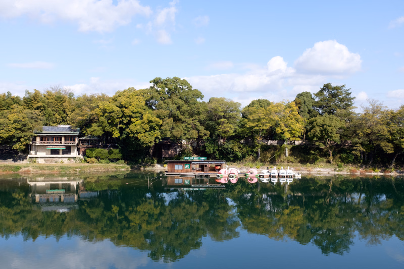 A calm lake reflects the surrounding lush green trees and a small boathouse under a partly cloudy blue sky.