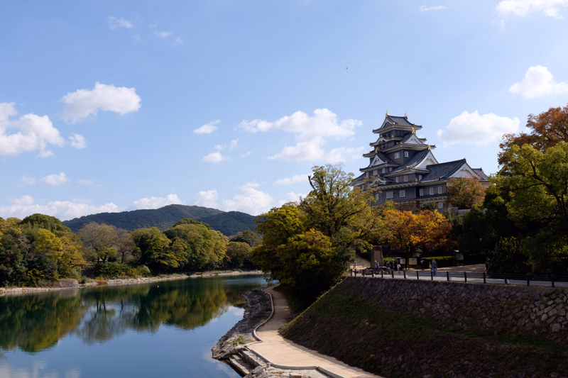 A traditional Japanese castle stands near a calm pond under a bright blue sky with scattered white clouds.