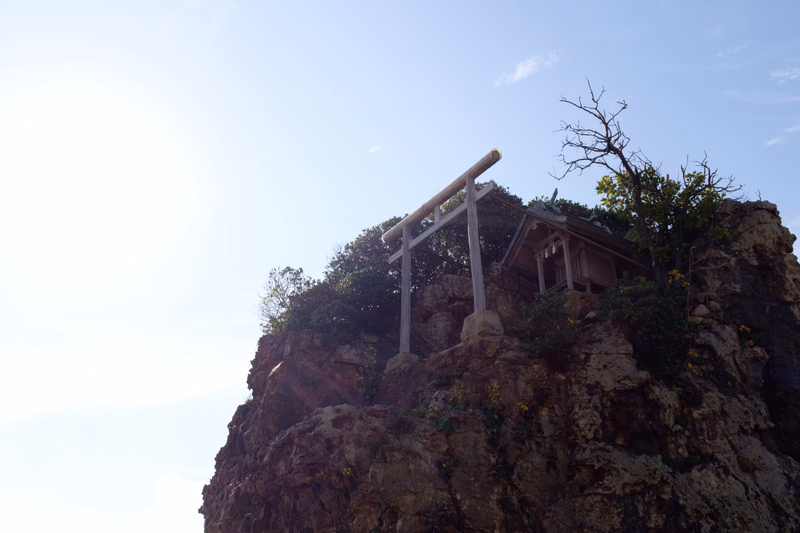 A wooden shrine structure is built precariously on the edge of a steep, rocky cliff under a bright sky.