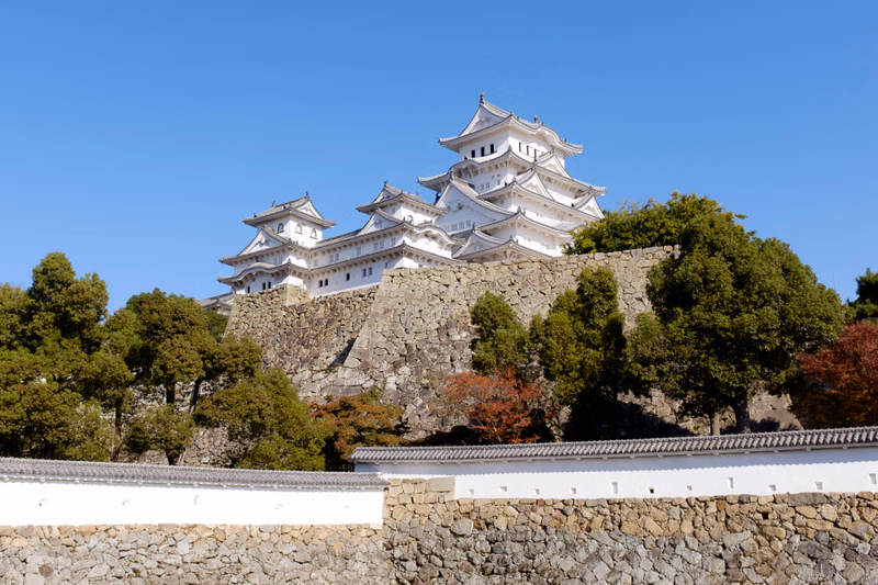 A white Japanese castle with multiple tiered roofs sits atop a stone hill under a clear blue sky, surrounded by trees and a lower stone wall.