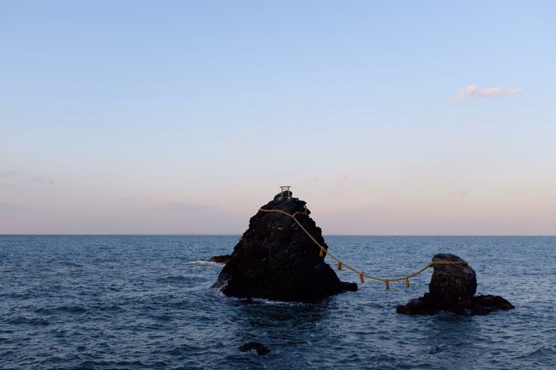 A small rock in the sea has a simple wooden structure on top, connected by a rope to another rock.