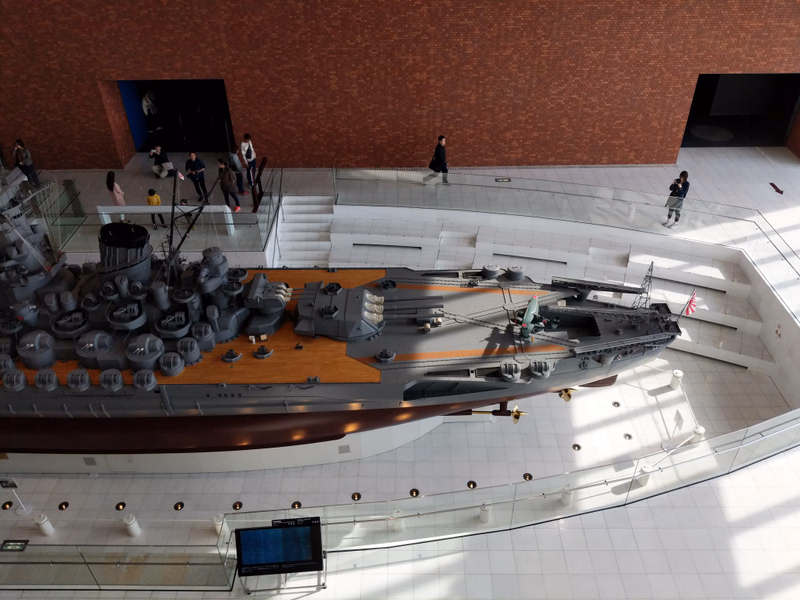 A large model of a warship is displayed inside a modern museum hall with high ceilings and brick walls.