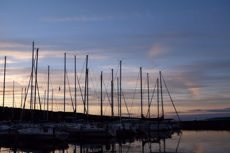 A silhouette of numerous sailboats moored in a harbor against a colorful twilight sky at sunset.