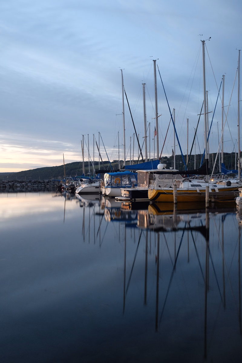 A group of sailboats are moored in a calm harbor at dusk, their masts reflecting clearly on the still water.