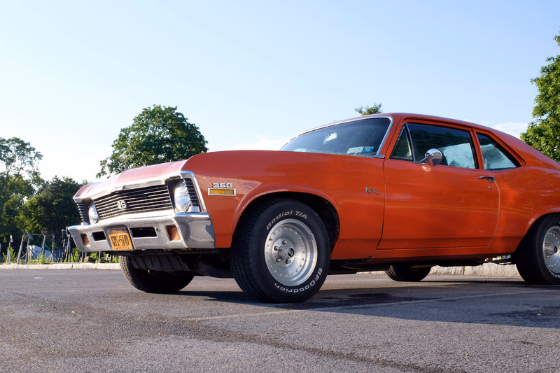 A bright orange 1960s Ford Fairlane muscle car is parked on asphalt under a clear sky.