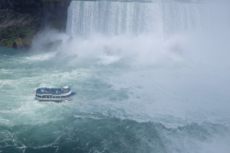 A tour boat navigates the turbulent water near the base of a large, powerful waterfall, creating significant spray.