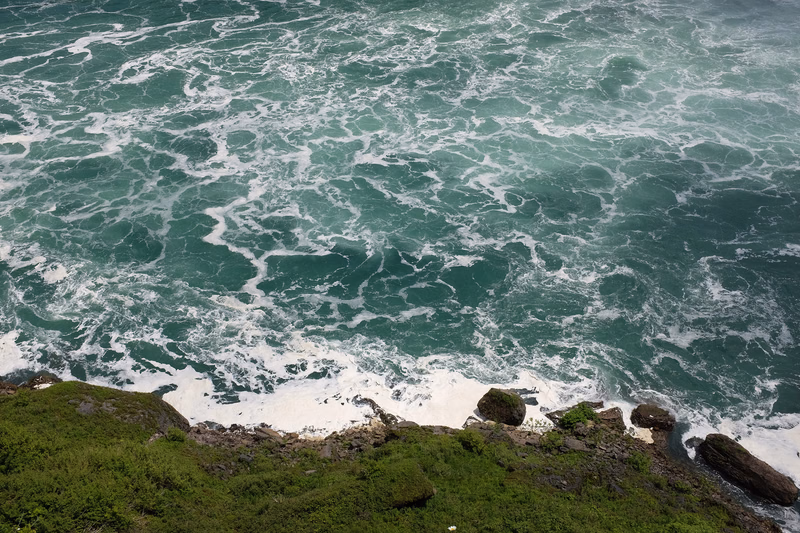 Rough turquoise ocean waves crash against a rocky coastline covered with green vegetation.