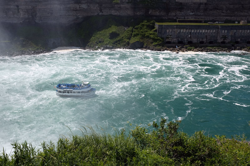 A tour boat navigates the turbulent, turquoise water near a large dam and waterfall.