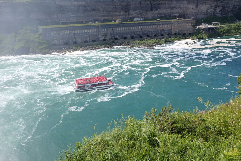 A tour boat navigates the turbulent, turquoise water below a large concrete dam structure surrounded by green grass.