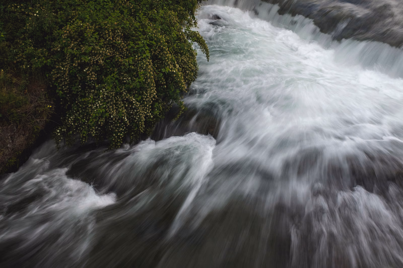 A fast-flowing, white waterfall cascades over dark rocks next to dense green foliage.