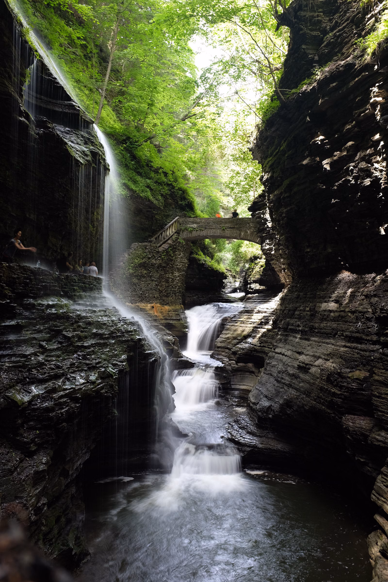 A multi-tiered waterfall cascades through a lush green forest, with a small wooden bridge crossing over the stream.