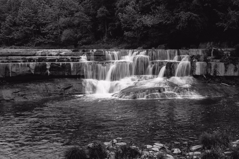 A wide view of a tiered waterfall cascading over rock ledges into a flowing river surrounded by dense trees.