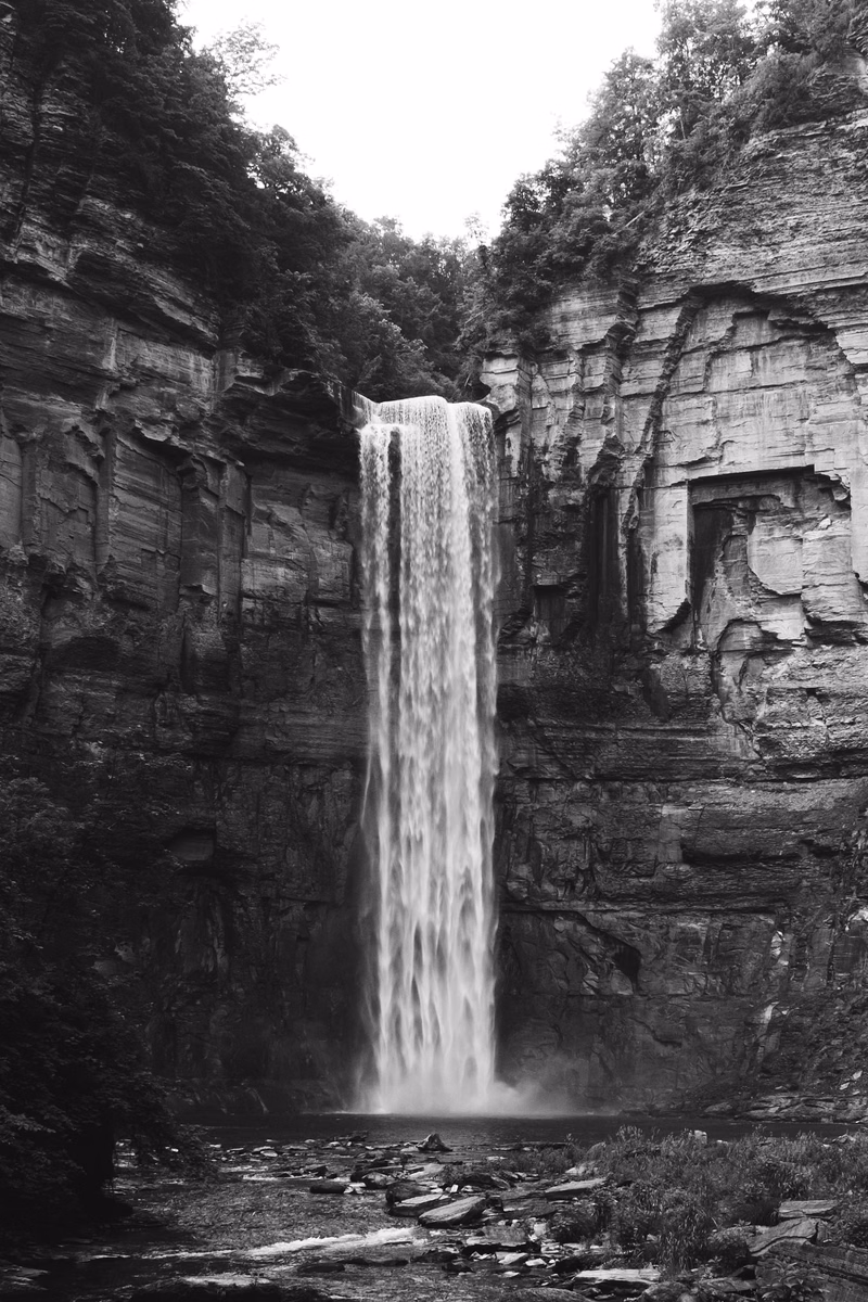 A tall, powerful waterfall cascades down a sheer, layered cliff face surrounded by dense forest.