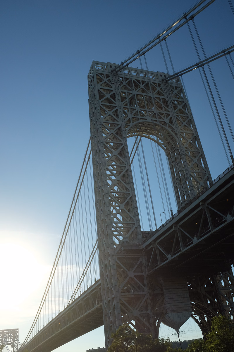 A large steel suspension bridge with a prominent arch rises against a clear blue sky.