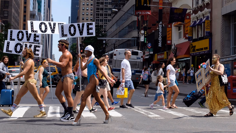 A group of people crossing a street in New York City, holding signs that read 'LOVE LOVE LOVE' and 'LOVE LOVE LOVE.' The scene is vibrant and full of energy.