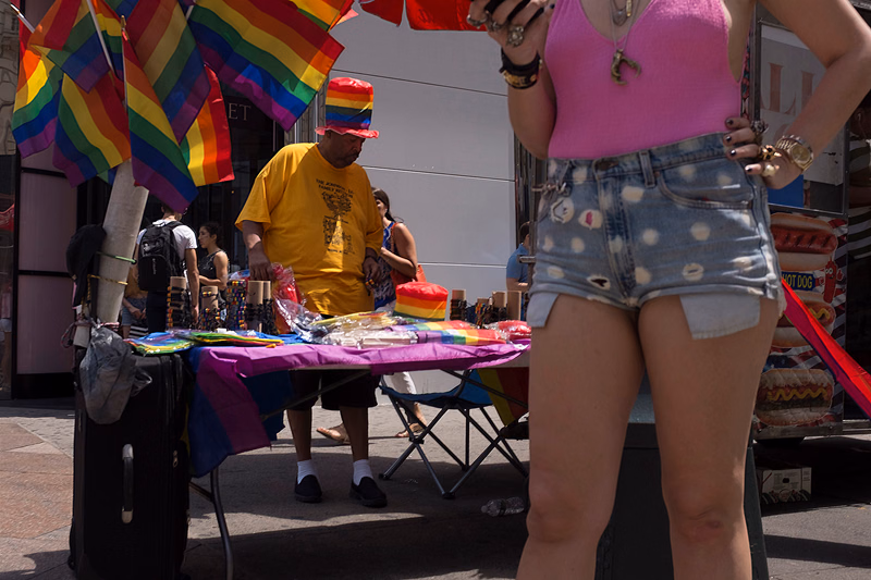 A person in a pink bikini and denim shorts stands near a vendor stall decorated with rainbow flags on a city street.