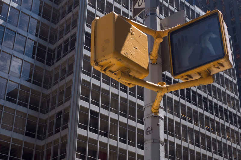 A yellow security camera is mounted on a pole in front of a modern glass skyscraper in a city setting.