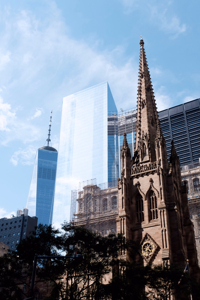 A tall, ornate gothic church stands in the foreground against a backdrop of modern glass skyscrapers under a partly cloudy sky.