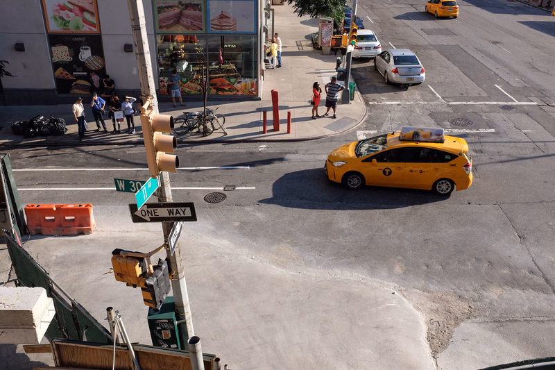 A yellow taxi is driving on a city street with a "ONE WAY" sign visible near a storefront displaying "cookin'".