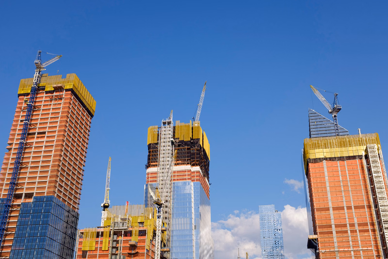 Several tall buildings under construction with visible cranes against a clear blue sky.