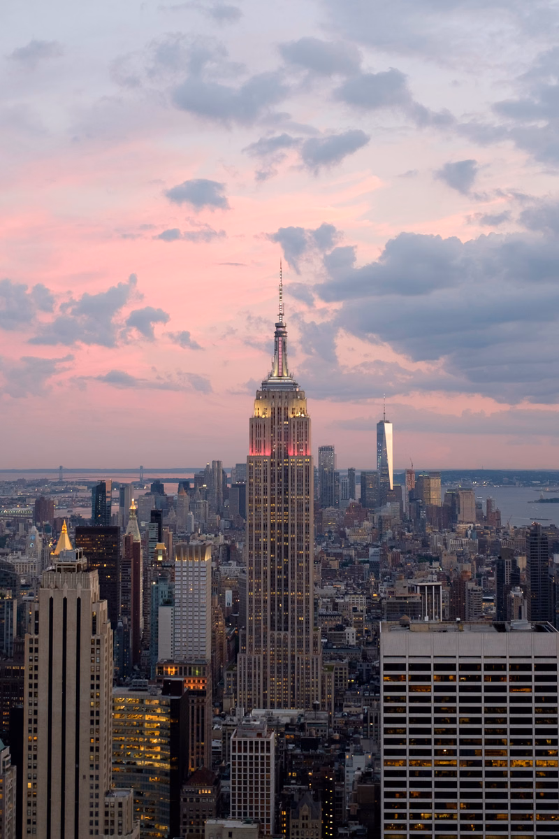 A tall skyscraper dominates the cityscape under a colorful sunset sky, surrounded by other high-rise buildings.