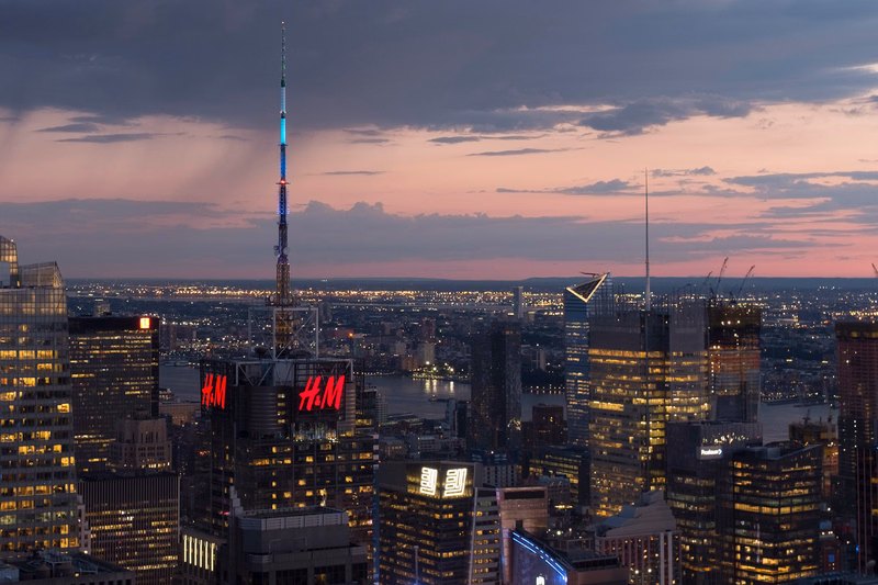 A tall communications tower stands over a dense cityscape at dusk, with several buildings displaying "H&M" signage.