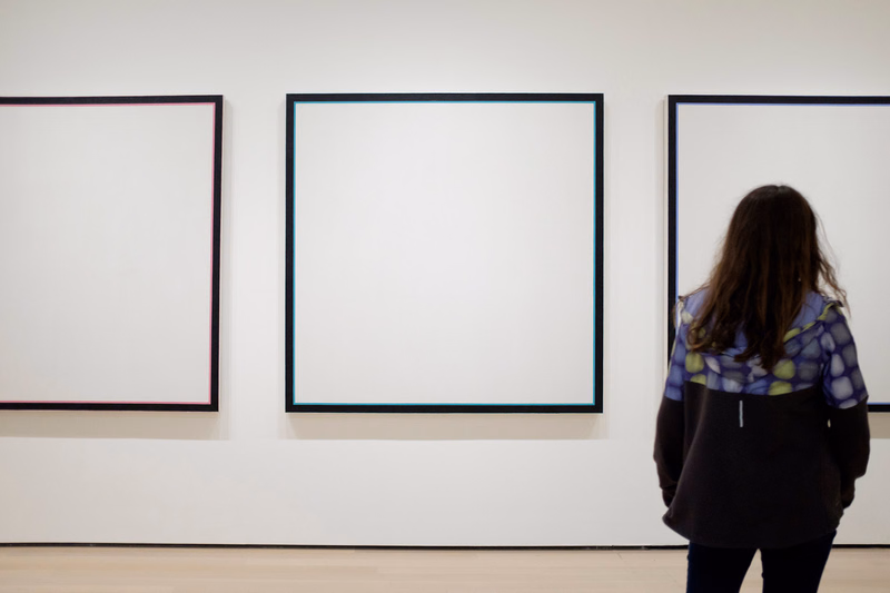 A woman stands in a bright art gallery viewing three large, framed white canvases on the wall.