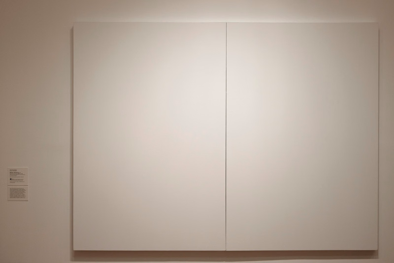 A large, minimalist abstract painting featuring two vertical panels of solid, light beige color displayed on a gallery wall.