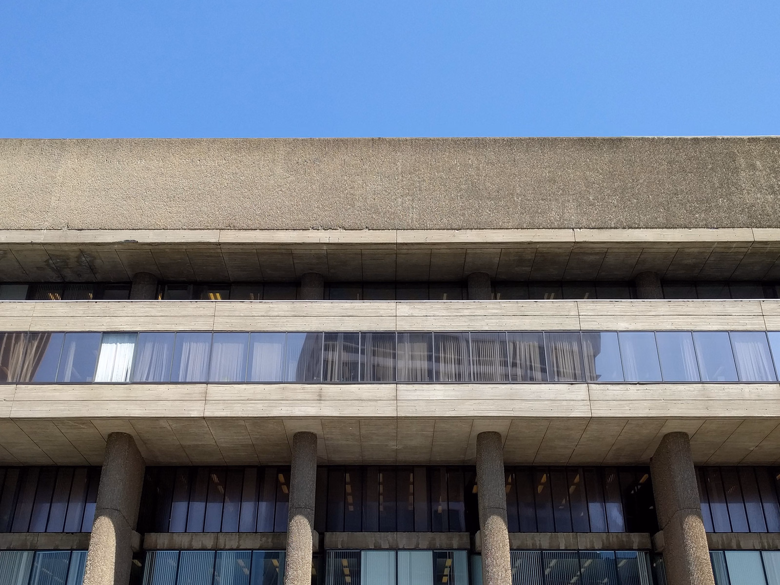 A modern building facade featuring multiple horizontal bands of windows set into textured concrete, set against a clear blue sky.