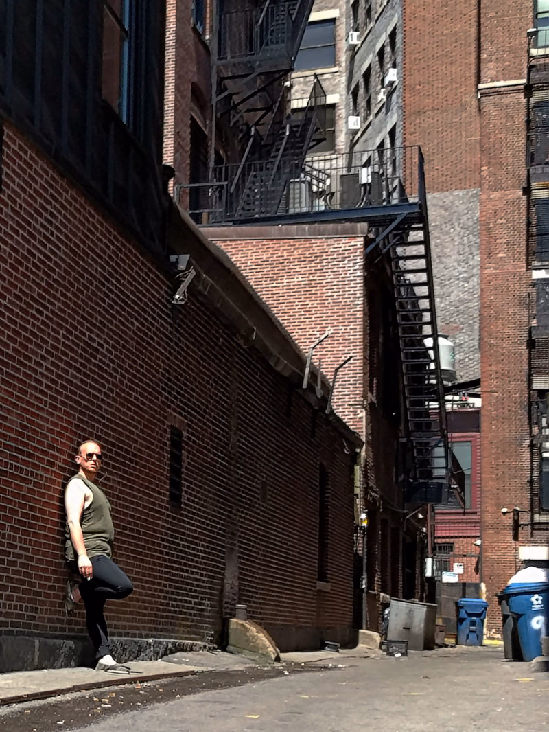 A man in a green shirt stands on a sidewalk next to a tall brick building with external fire escapes.