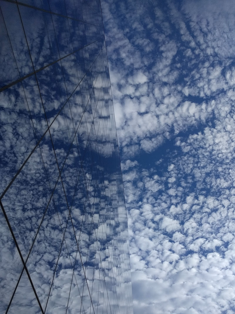 A modern glass building reflects a bright blue sky filled with scattered white cumulus clouds.