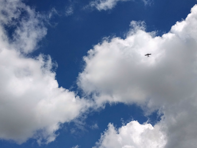 A small airplane flies across a bright blue sky dotted with white cumulus clouds.