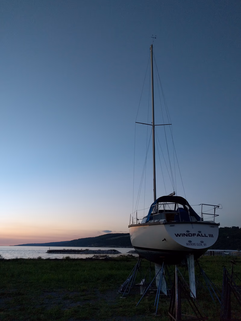 A sailboat is seen on a grassy shore near a body of water, with a beautiful sunset in the background.