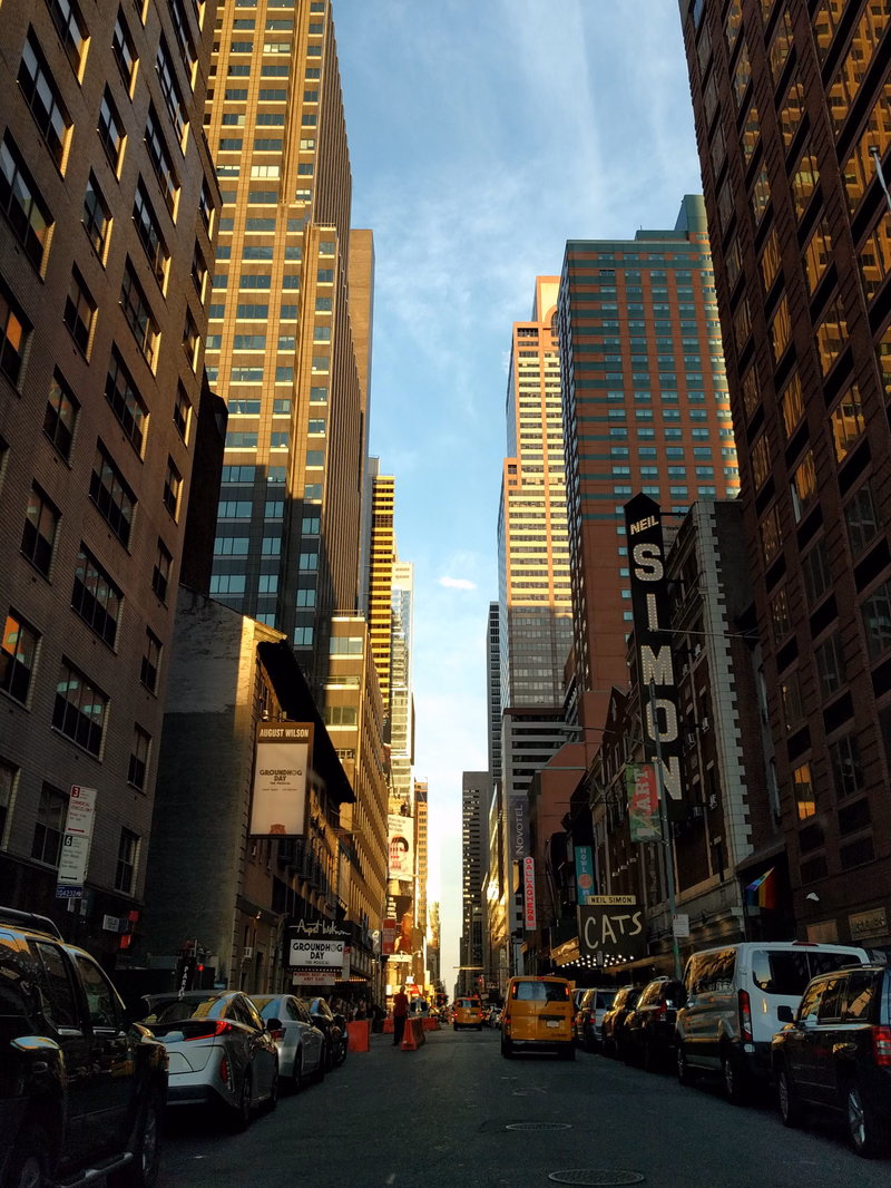 A view down a city street lined with tall skyscrapers, featuring a prominent "NEIL SIMON" sign on a building.