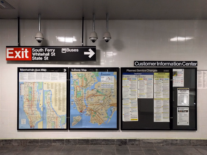 A set of transit maps and information boards in a subway station, including signs for "Exit South Ferry Whitehall St State St" and "Manhattan Bus Map".