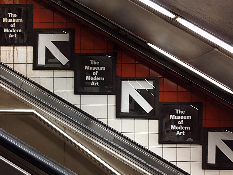 A series of black signs with white arrows and the text "The Museum of Modern Art" are mounted on a red tiled wall next to an escalator.