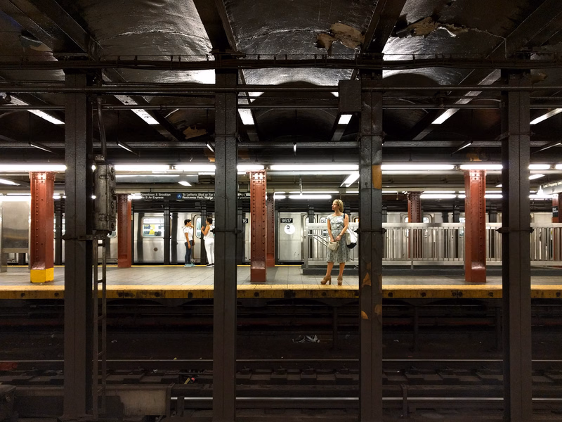 A woman stands on a subway platform under a low, industrial ceiling, with train tracks visible in the foreground.