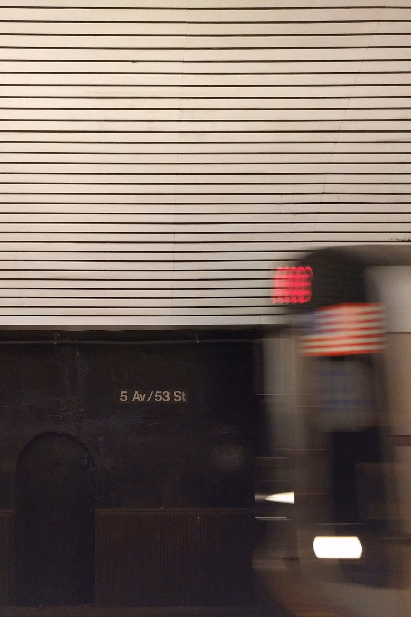 A subway train is visible behind a wall with horizontal slats, displaying a sign that reads "5 Av / 53 St".