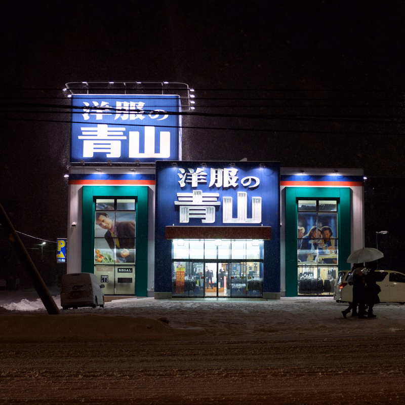 A brightly lit store at night with a large sign reading "洋服の青山" (Yōfuku no Aoyama) above the entrance.