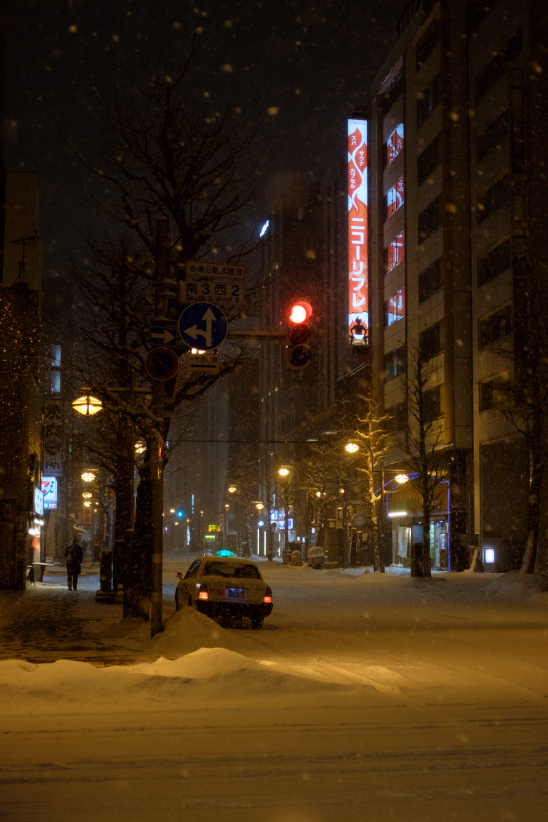 A snowy night scene on a city street with illuminated buildings and a traffic light displaying red.