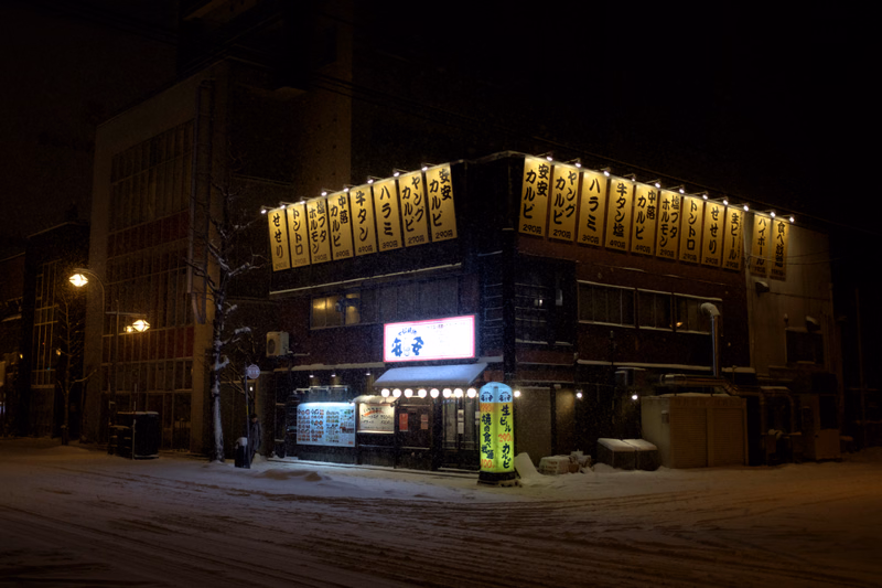 A Japanese restaurant at night in the snow, with a prominent illuminated sign that reads "名菜".