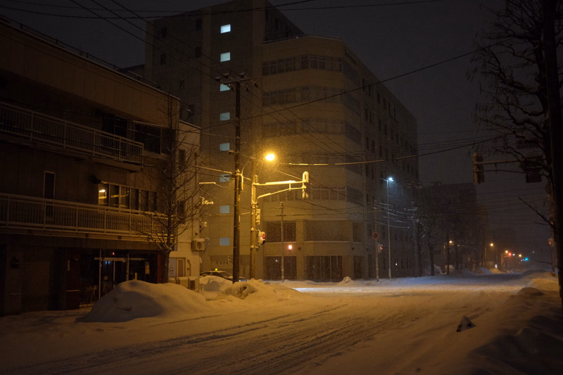 A snowy city street at night with illuminated buildings and streetlights.