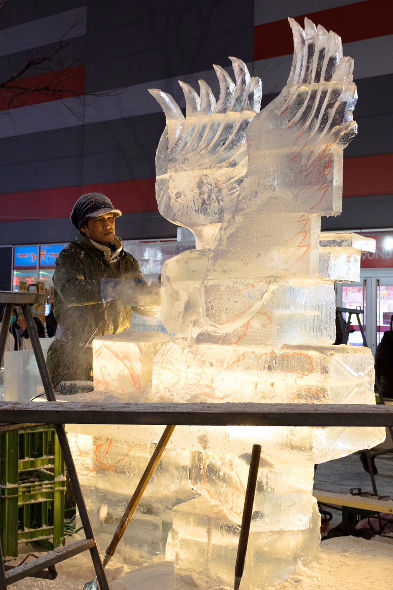 A man works on an intricately carved ice sculpture in a snowy environment.