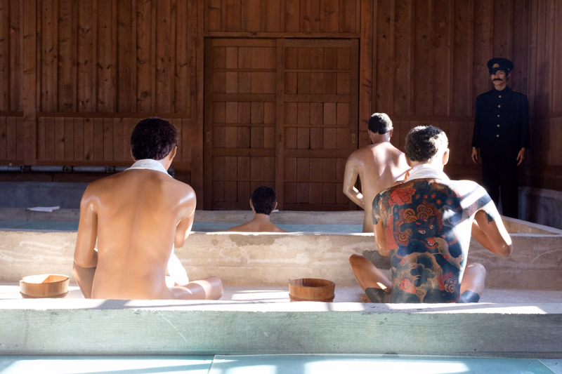 Several men with tattoos are sitting in a wooden bathing area, while a man in a dark uniform stands nearby.