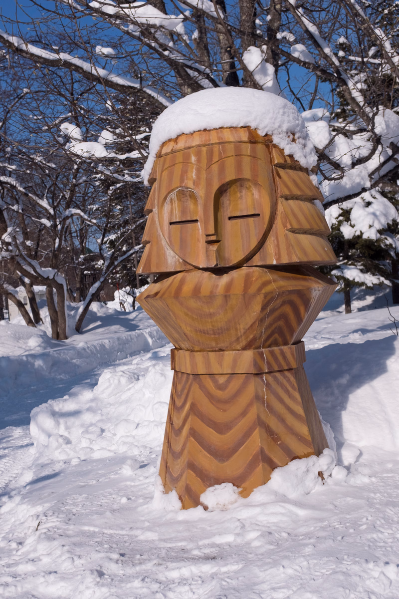 A large, wooden, carved sculpture resembling a snowman stands in the snow outdoors under bare trees.