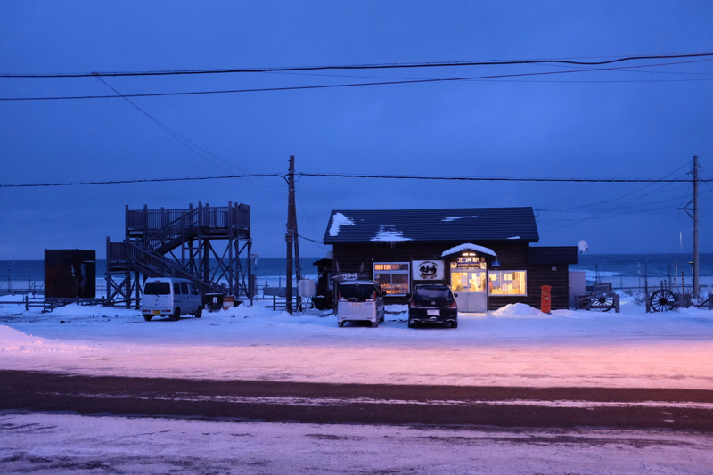 A small beach hut with illuminated windows sits near a wooden pier on a snowy evening by the ocean.