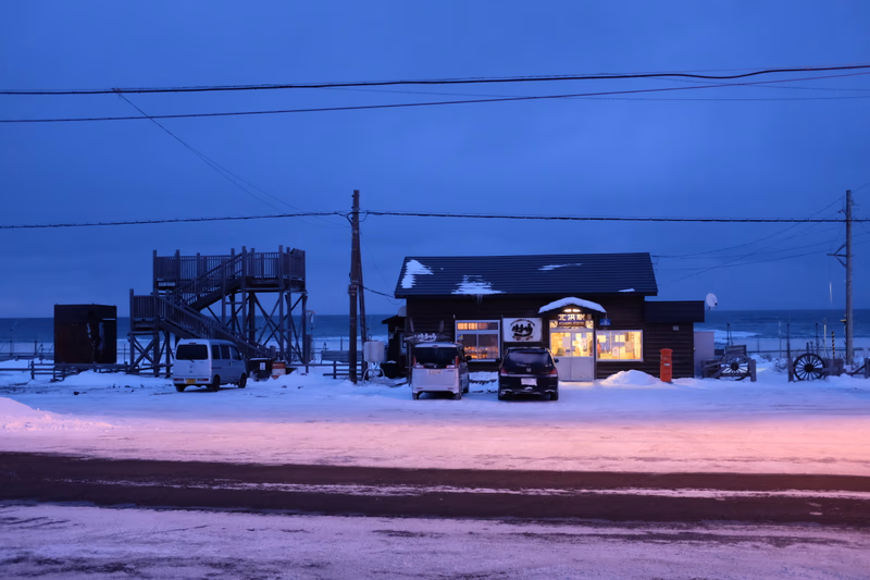 A small building with a snow-covered roof, a truck parked in front, and a road leading to a snowy landscape with a body of water in the background.