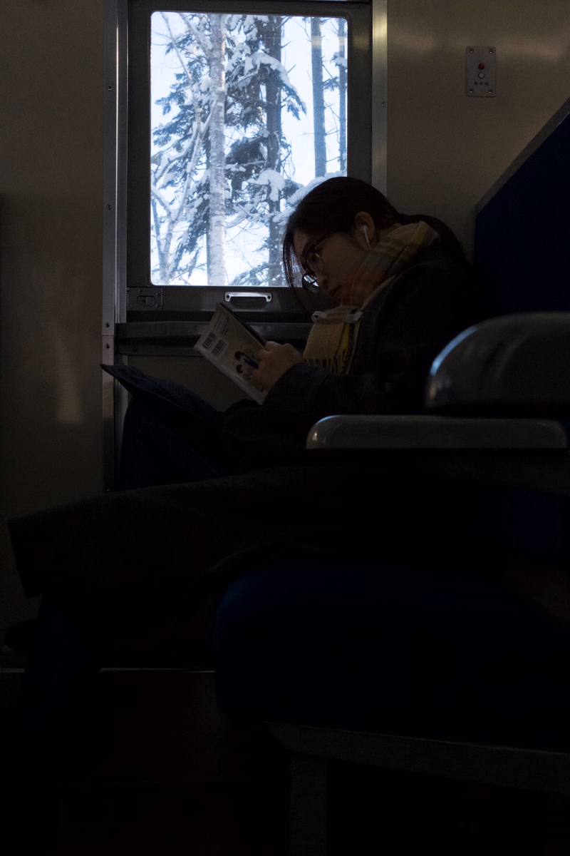 A person wearing glasses reads a book while sitting indoors near a window showing trees outside.