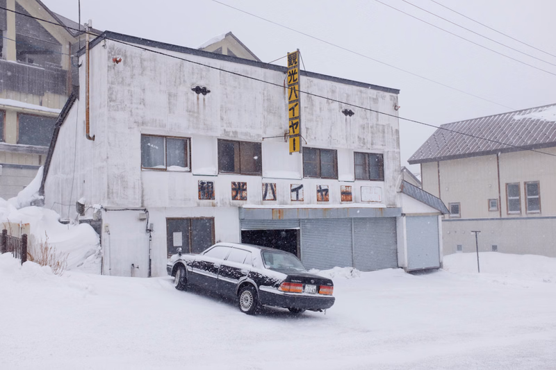A white building with a vertical sign reading "光ハッチャ" is covered in snow, and a black car is parked in the snowy foreground.
