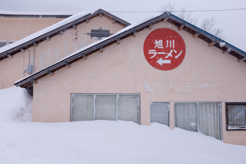 A small ramen shop building covered in snow features a circular sign that reads "旭川ラーメン" (Asahikawa Ramen).
